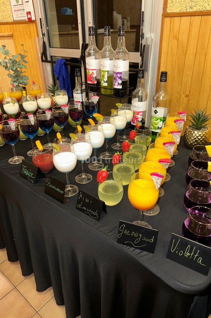Table avec divers cocktails colorés, étiquetés, décorés de fruits ou plantes, disposés sur une nappe noire.