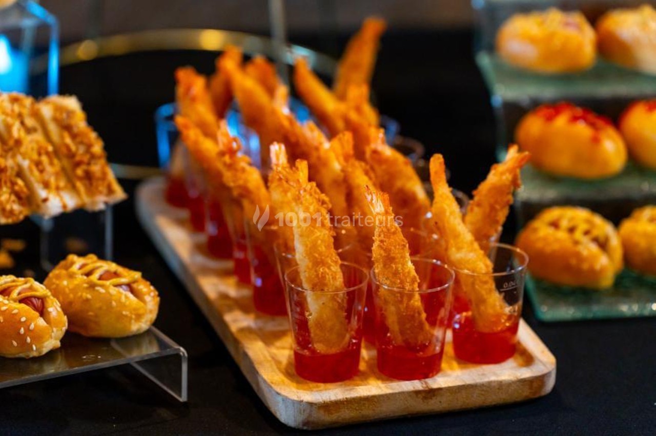 Crevettes panées présentées dans des verrines de sauce rouge, entourées de petits pains garnis sur une table.
