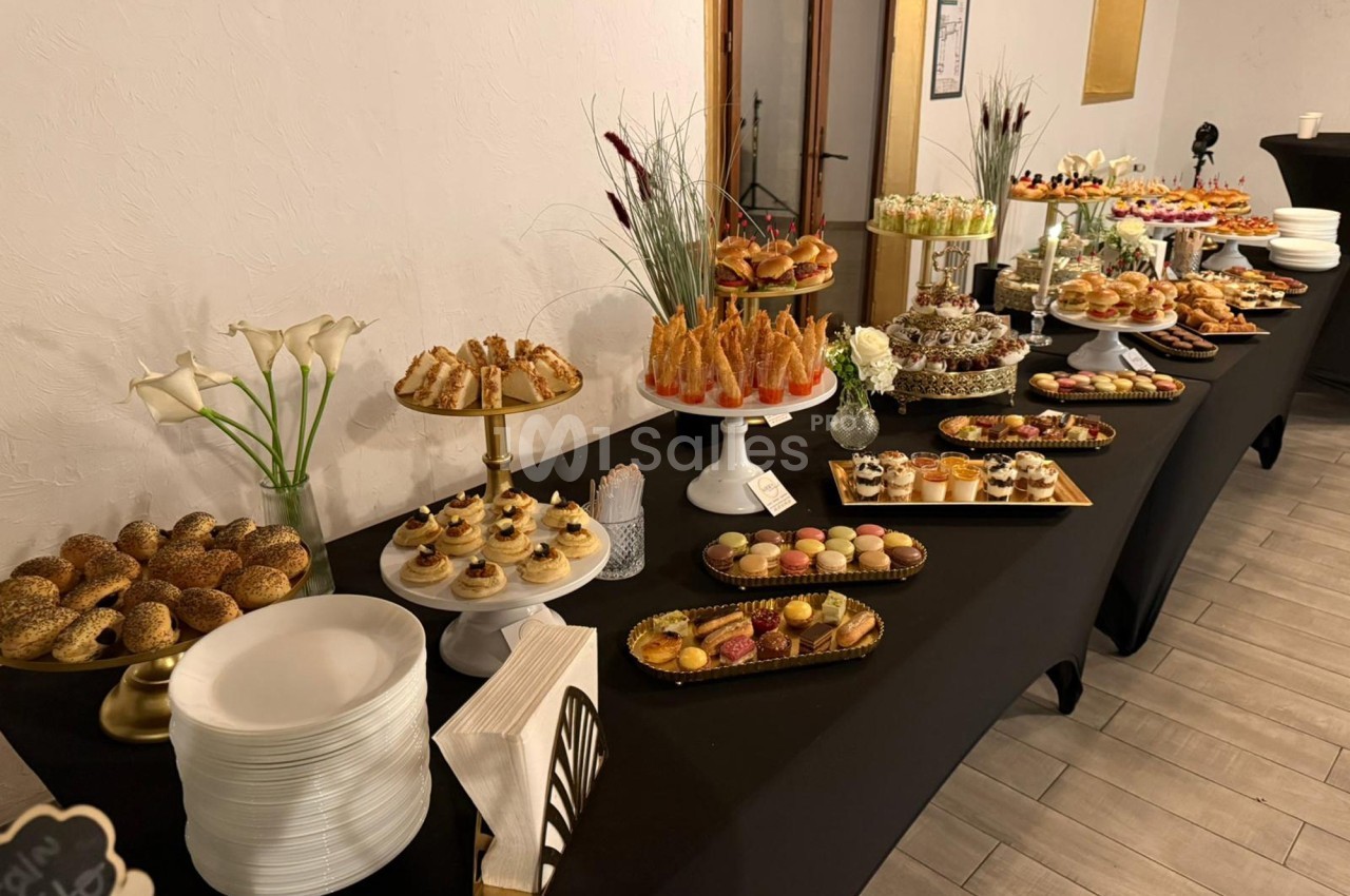 Buffet varié avec pâtisseries, pains, verrines et assiettes disposés sur une table noire dans une salle lumineuse.