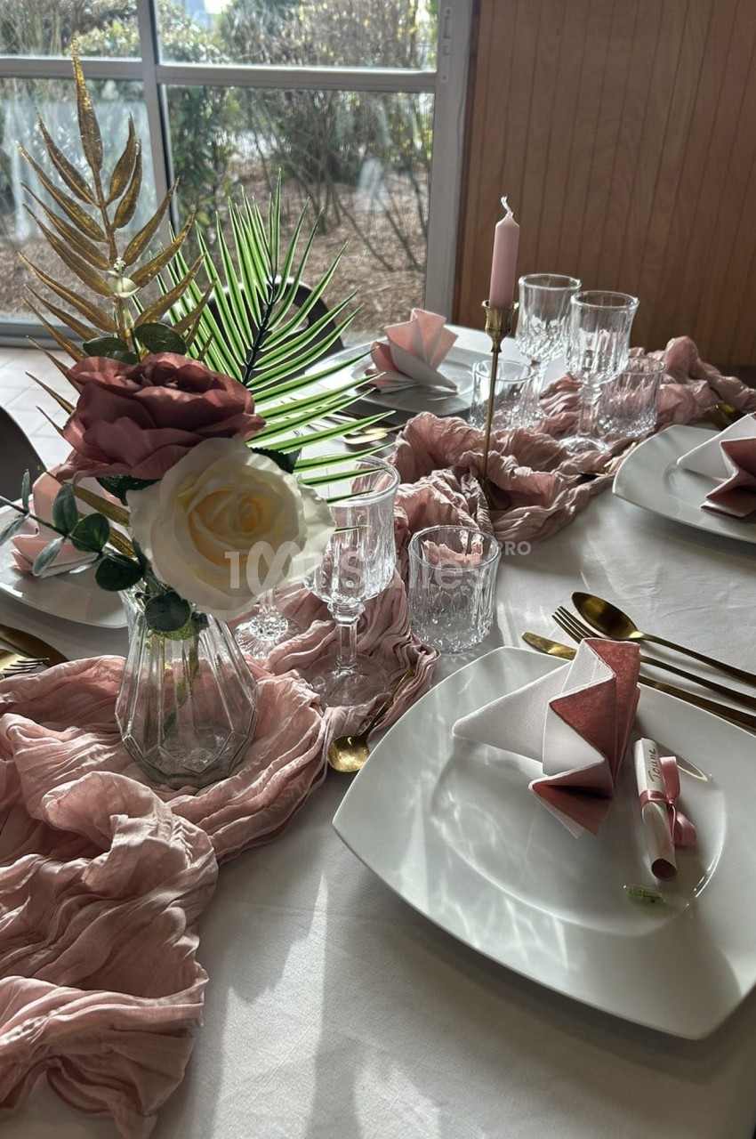 Table décorée avec des assiettes blanches, serviettes roses pliées, verres, bougies et un bouquet de fleurs dans un vase.