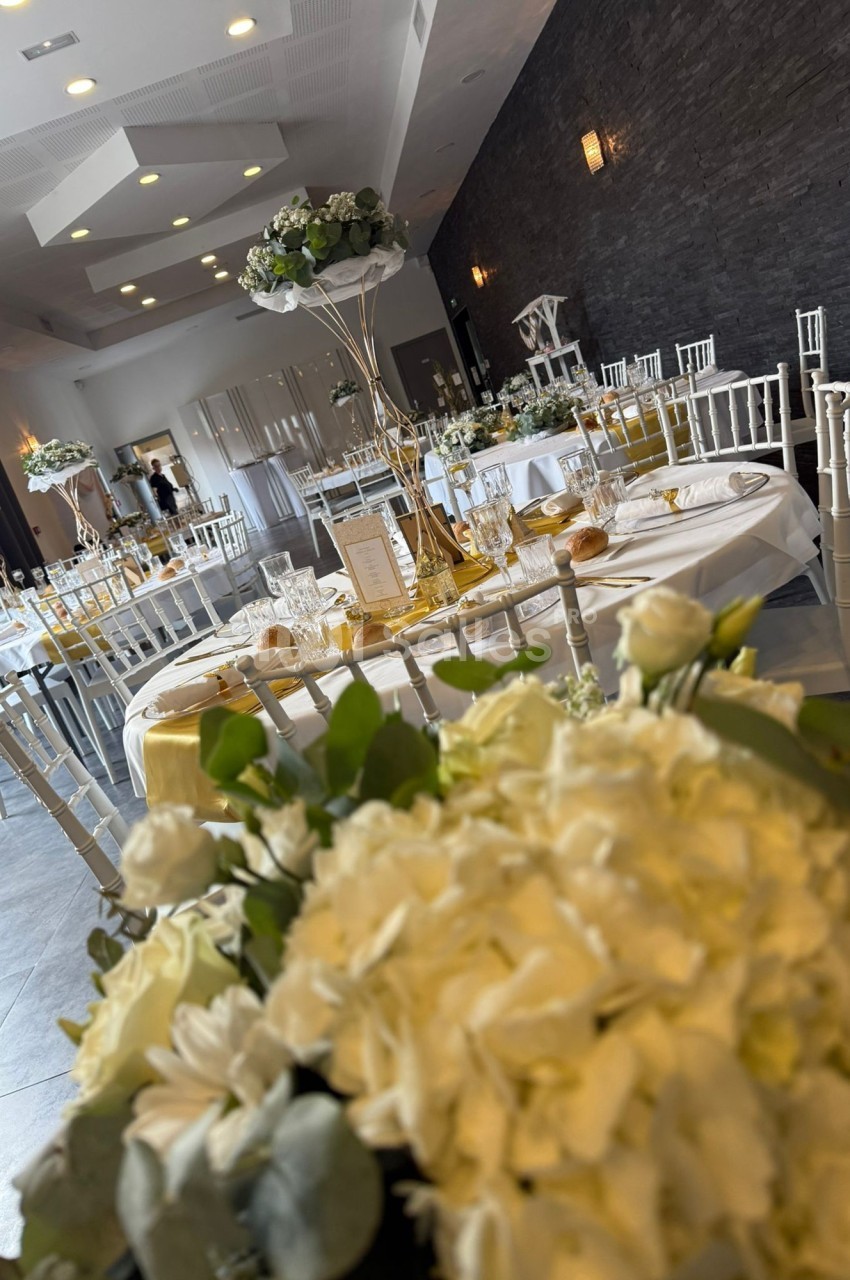 Salle de réception élégamment décorée avec des tables dressées, des chaises blanches et des centres de table fleuris.