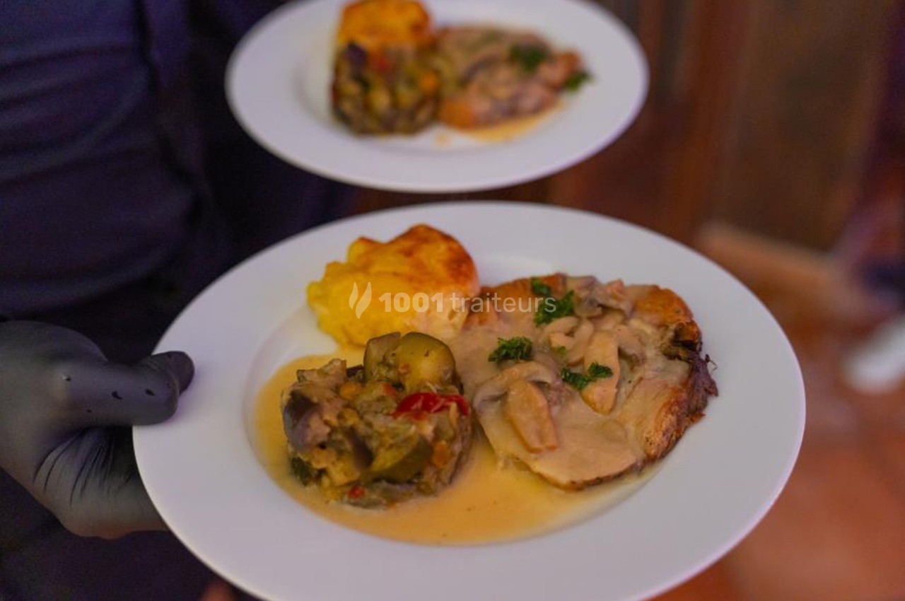 Deux assiettes contenant un plat de viande en sauce, légumes et gratin, servies par une personne portant des gants noirs.