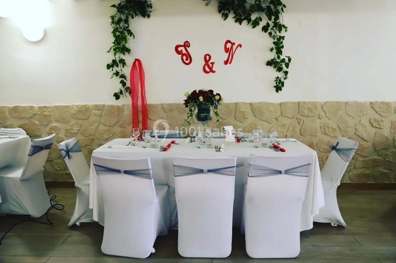Table décorée pour un événement avec nappes blanches, chaises ornées de rubans, fleurs au centre et initiales murales.