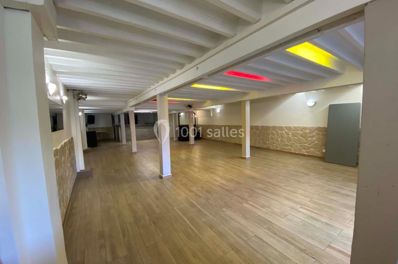Salle vide avec sol en parquet, murs en pierre et plafond blanc avec poutres apparentes et éclairage coloré.