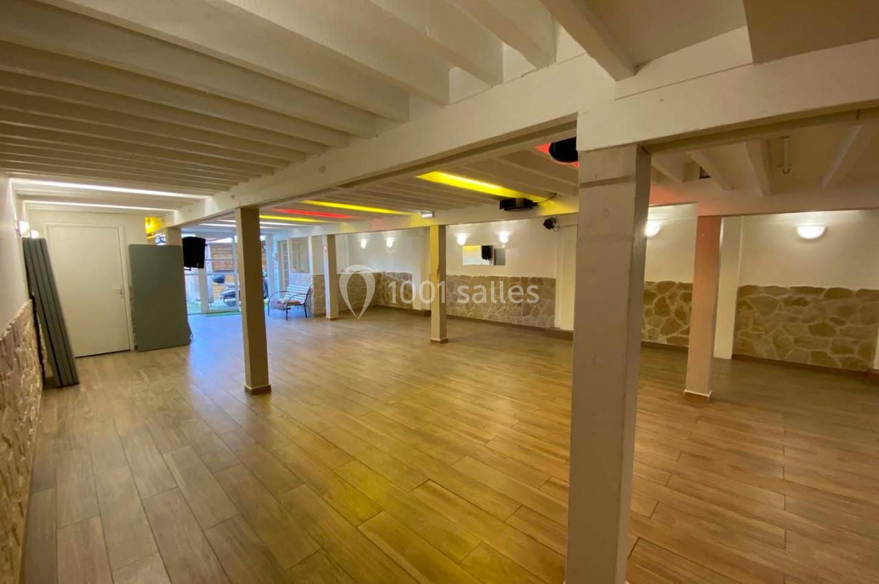 Salle spacieuse avec sol en parquet, murs en pierre et poutres blanches apparentes, éclairée par des lumières modernes.