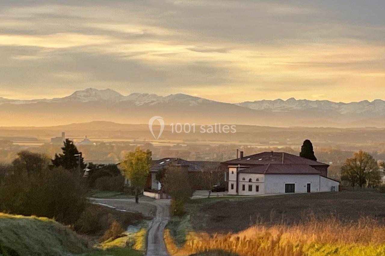 Chemin de campagne menant à des bâtiments entourés de champs, avec des montagnes et un ciel doré en arrière-plan.