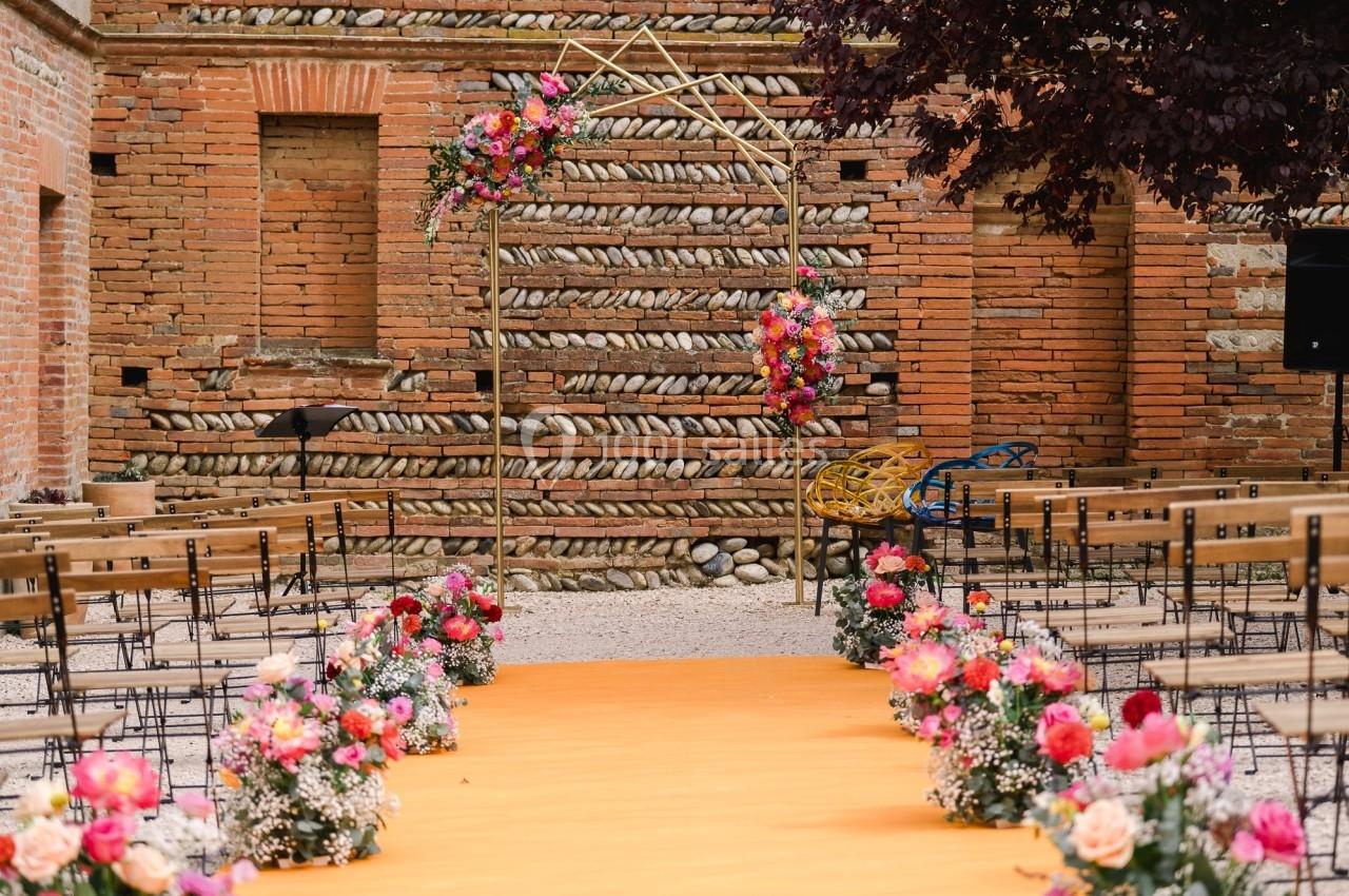 Allée décorée de fleurs menant à une arche ornée, installée pour une cérémonie en extérieur devant un mur en briques.