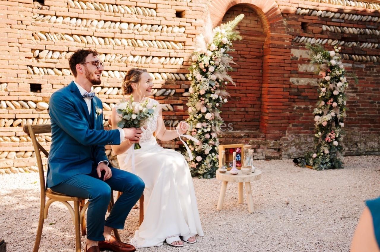 Un couple assis lors d'une cérémonie de mariage en extérieur, entouré de décorations florales et d'un mur en briques.