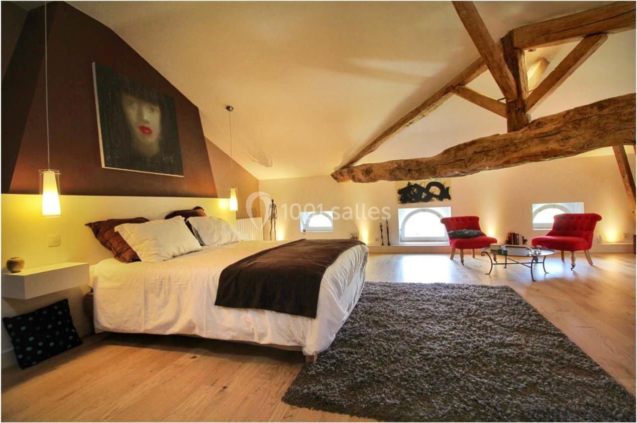 Chambre spacieuse sous combles avec lit double, poutres apparentes, fauteuils rouges et lumière naturelle.