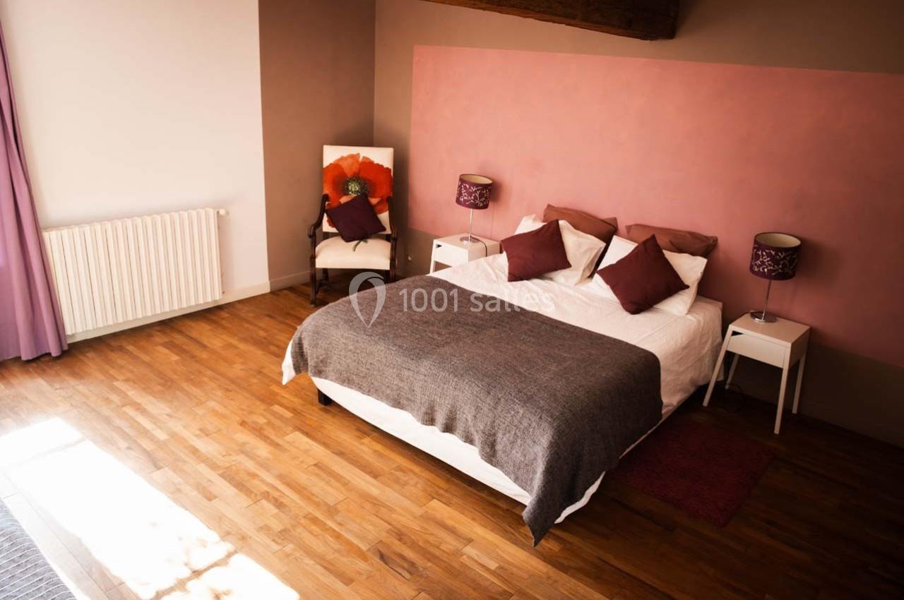 Chambre avec lit double, couvre-lit gris, coussins rouges, murs rose et beige, parquet et fauteuil avec coussin orange.