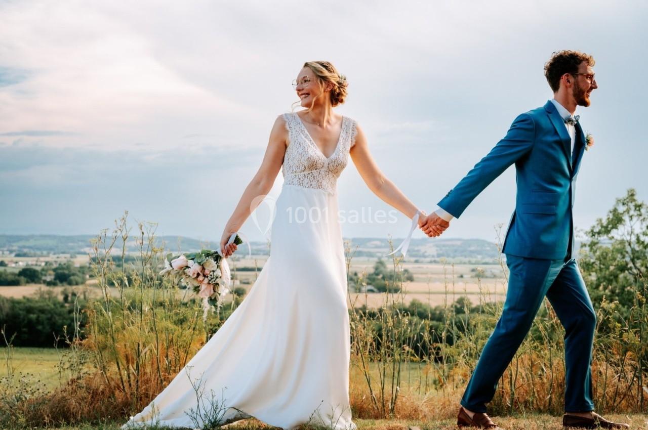Un couple en tenue de mariage marche main dans la main dans un paysage champêtre sous un ciel partiellement nuageux.