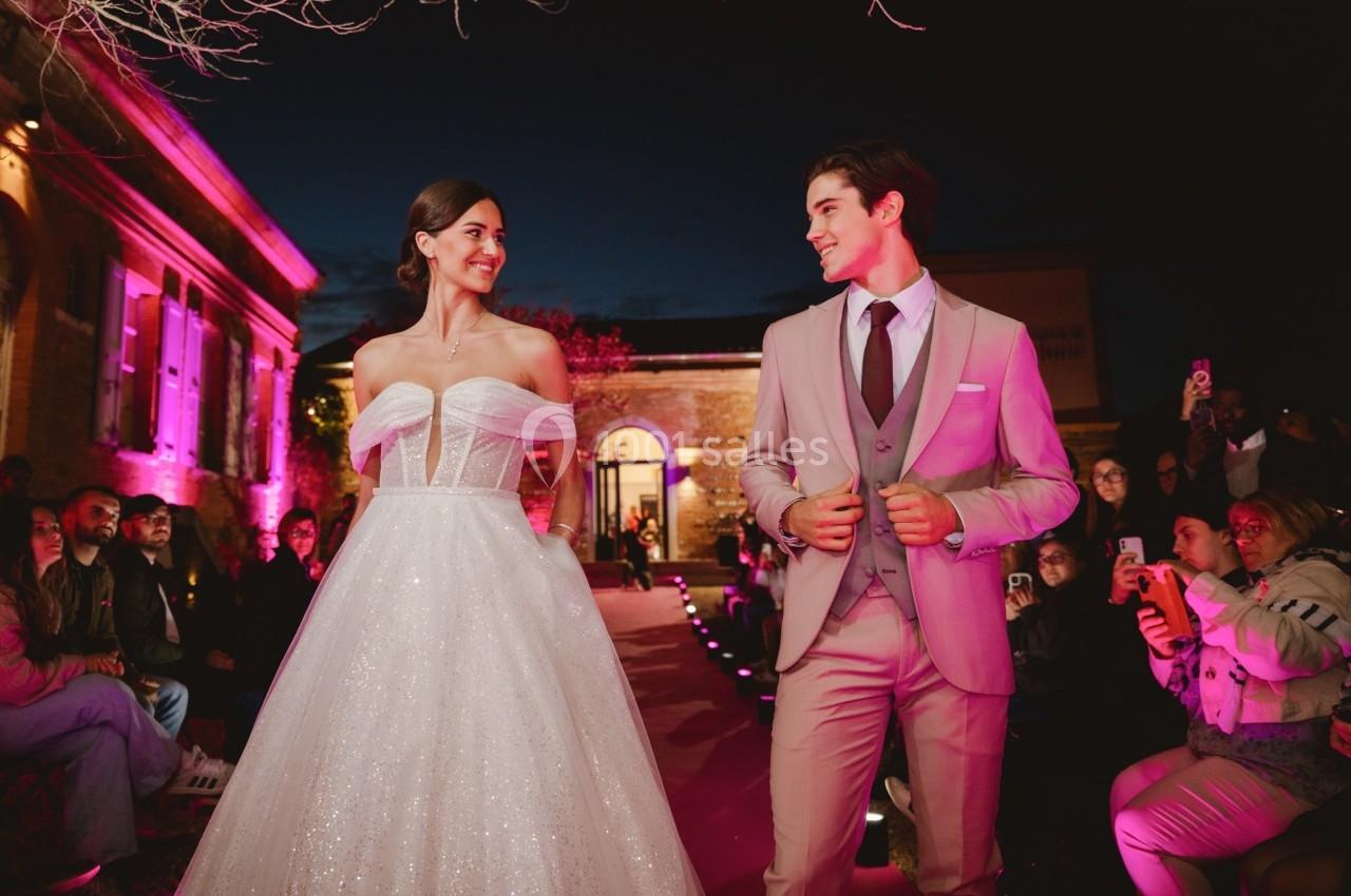 Un couple en tenue de mariage défile sur un podium extérieur éclairé par des lumières roses, devant un public.