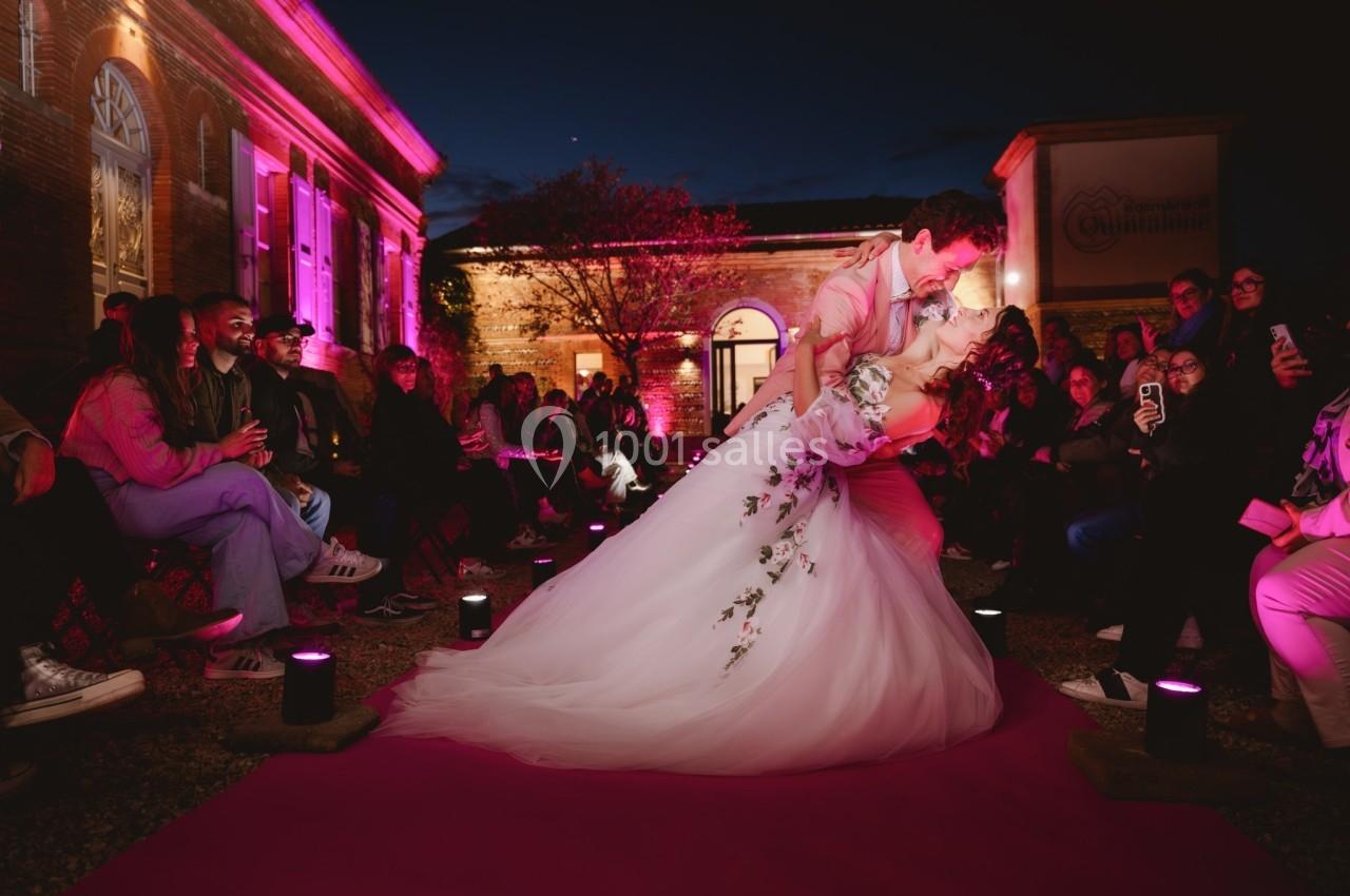 Un couple de mariés danse sur un tapis rose, entouré d'invités assis, dans une ambiance nocturne éclairée en rose.