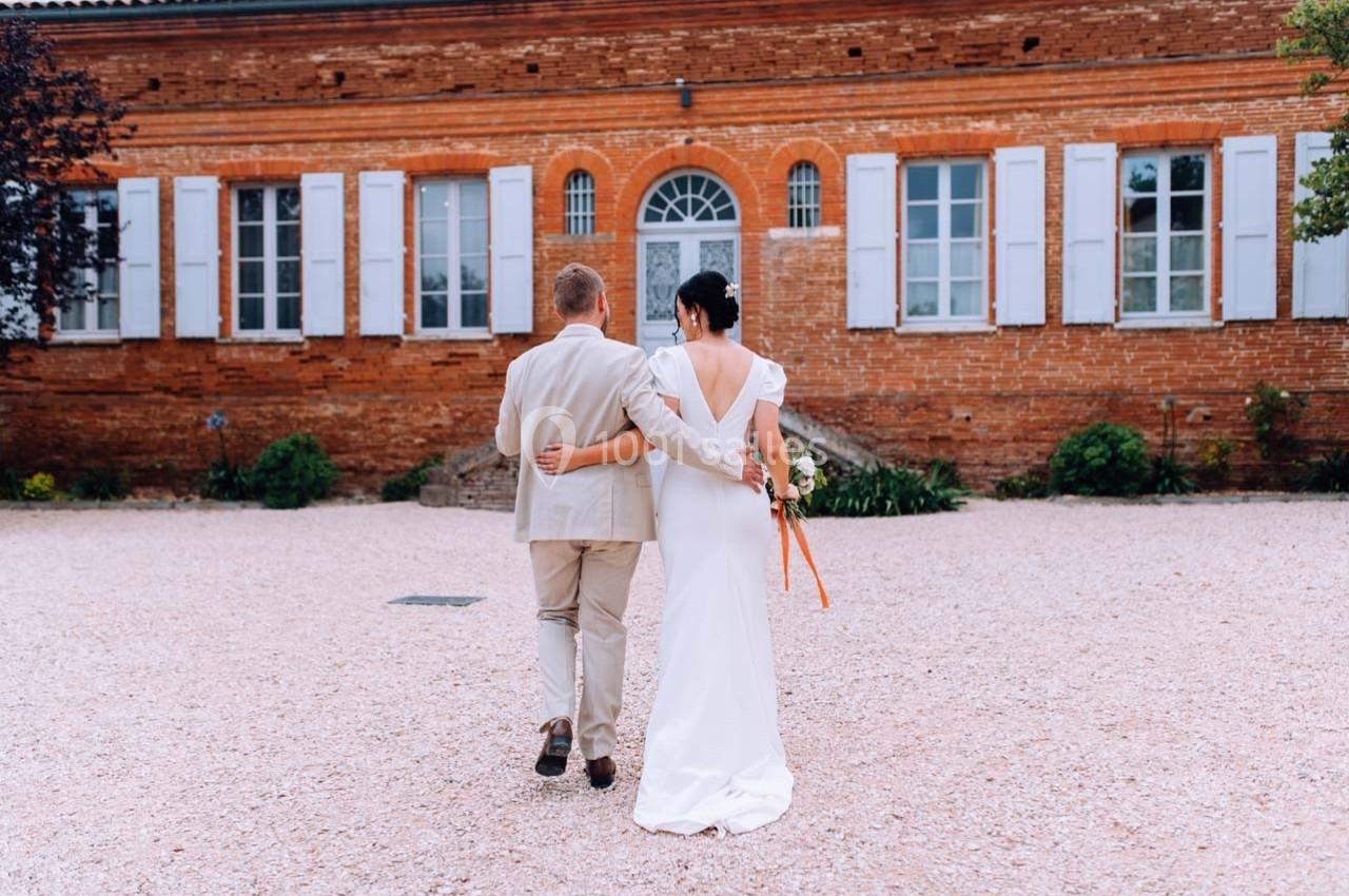Un couple de dos, marchant dans une cour en gravier devant un bâtiment en briques rouges avec des volets blancs.