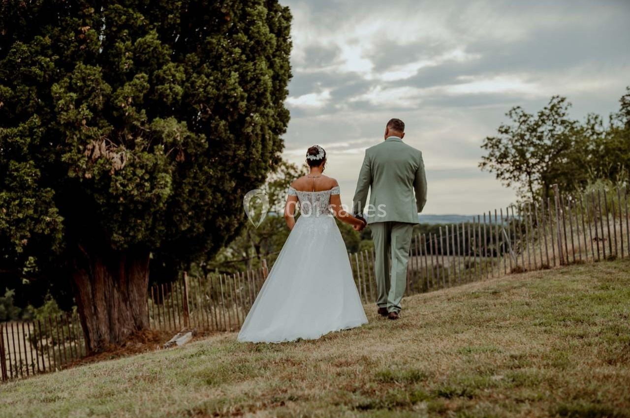 Un couple en tenue de mariage marche main dans la main sur une pelouse, entouré d'arbres et de clôtures en bois.