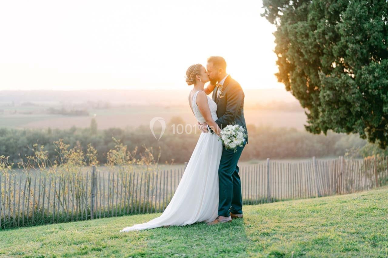 Un couple en tenue de mariage s'embrasse dans un jardin au coucher du soleil, près d'une clôture et d'un arbre.
