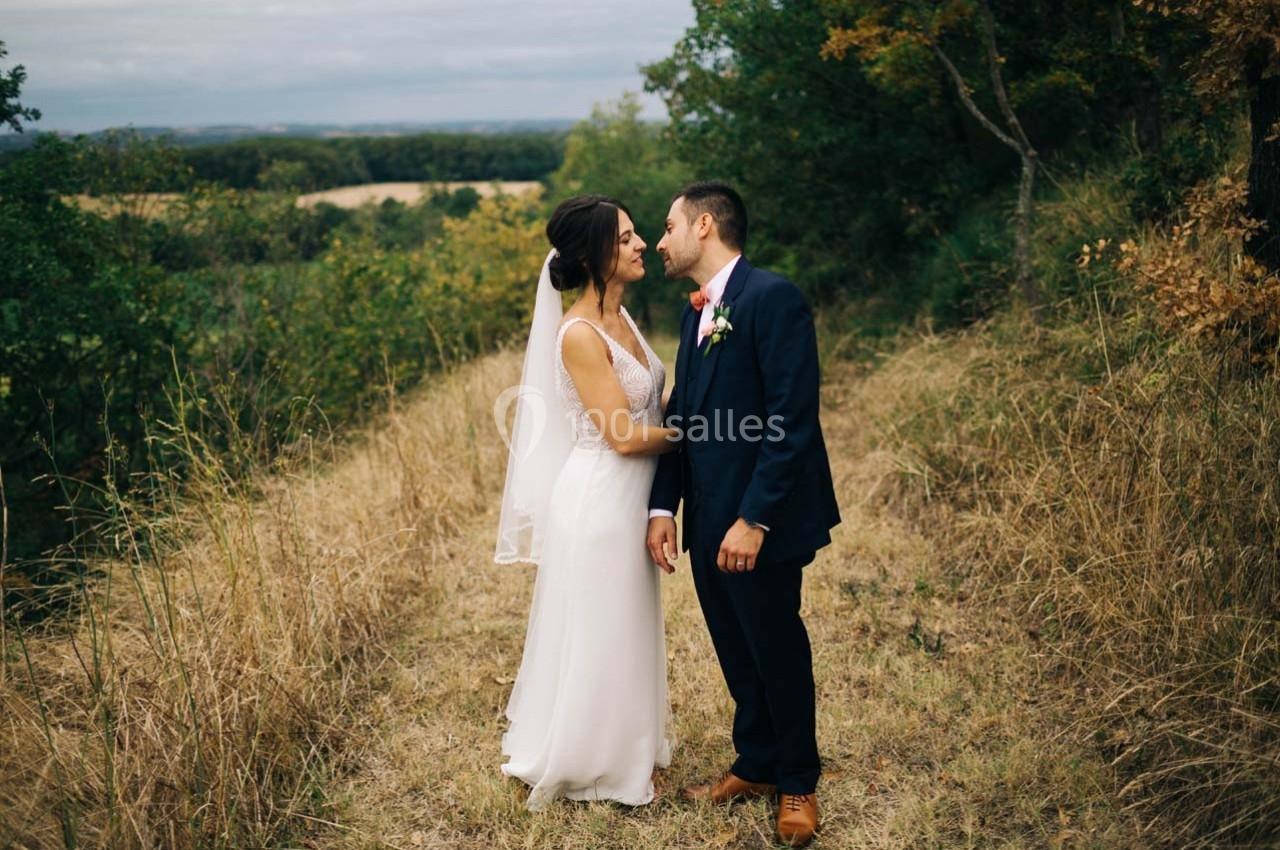 Un couple en tenue de mariage se tient sur un chemin herbeux entouré de végétation, échangeant un moment intime.