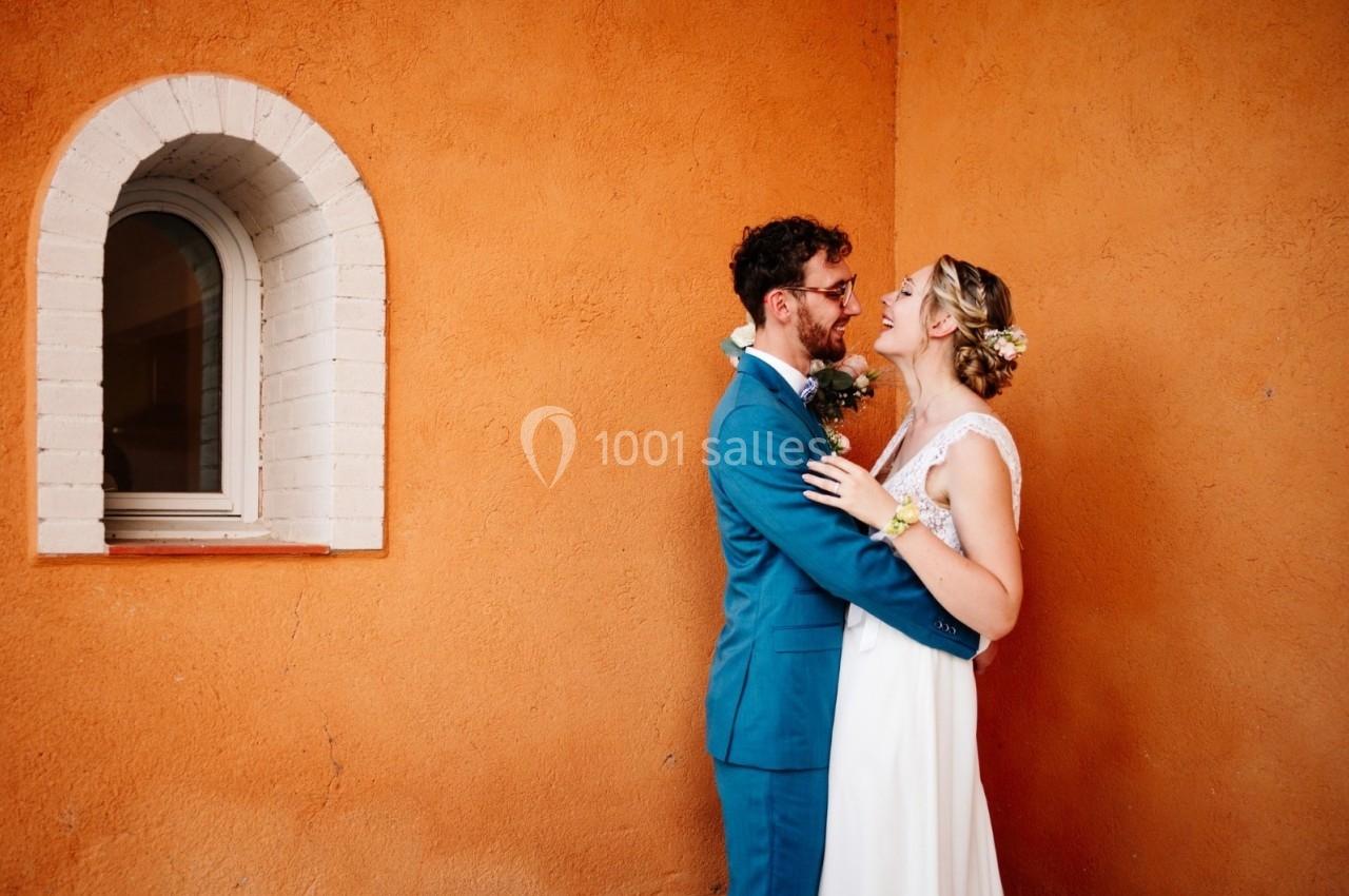 Un couple en tenue de mariage s'enlace devant un mur orange avec une petite fenêtre voûtée.