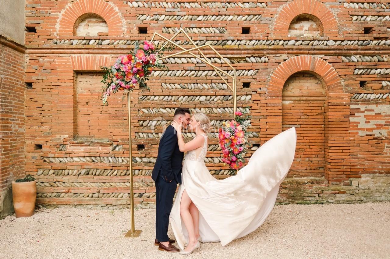 Un couple de mariés s'embrasse sous une arche décorée de fleurs colorées, devant un mur en briques anciennes.