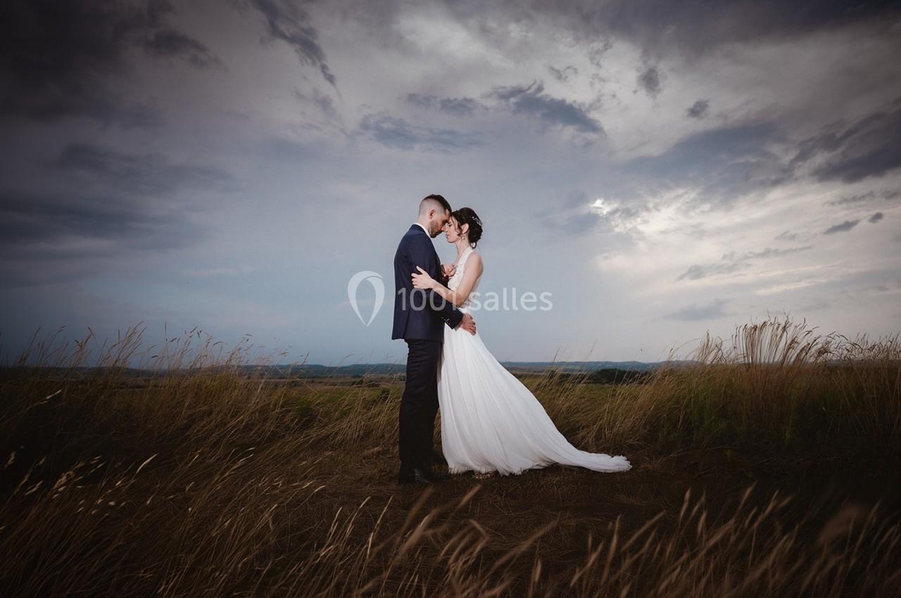 Un couple en tenue de mariage s'enlace dans un champ sous un ciel nuageux.
