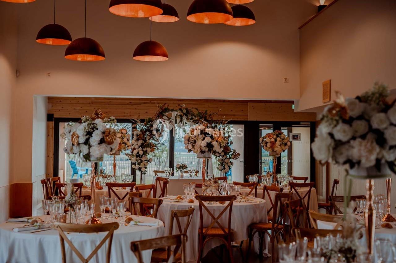 Salle de réception décorée pour un mariage avec des tables rondes, des chaises en bois et des compositions florales…