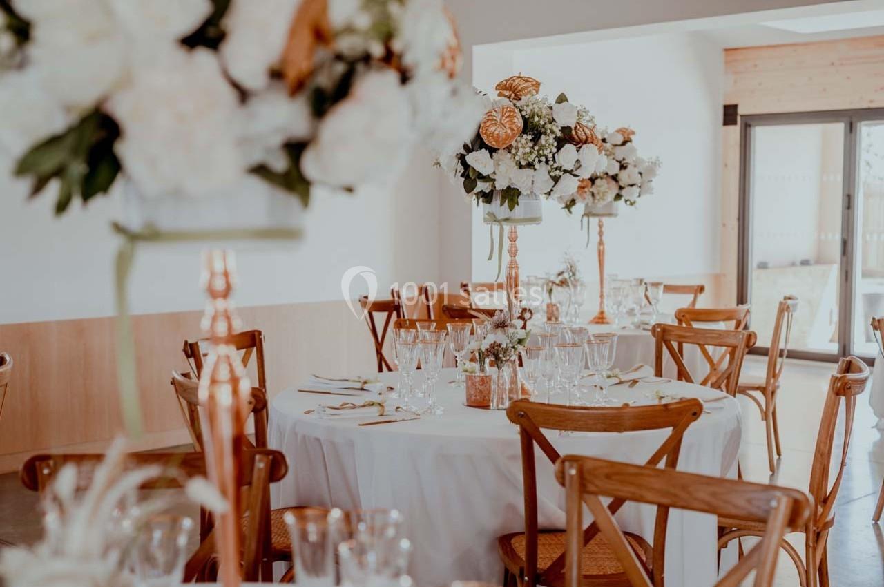 Salle décorée pour un événement avec tables rondes, nappes blanches, chaises en bois et centres de table fleuris.