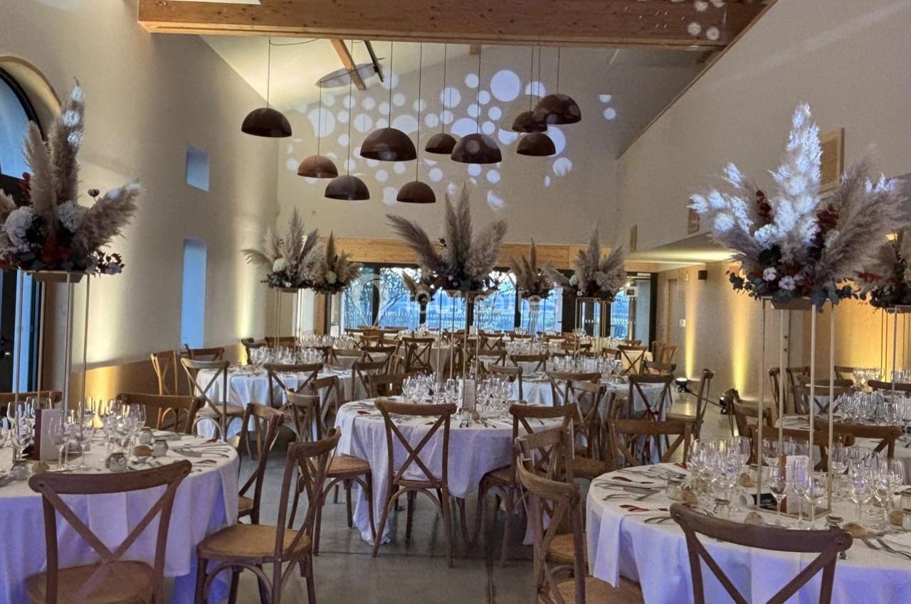 Salle de réception décorée avec des tables rondes dressées, des chaises en bois et des compositions florales suspendues.