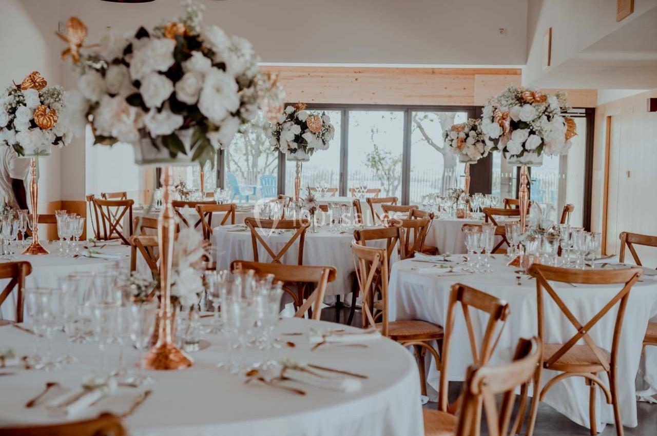 Salle de réception décorée avec des tables rondes, nappes blanches, chaises en bois et grands bouquets de fleurs.