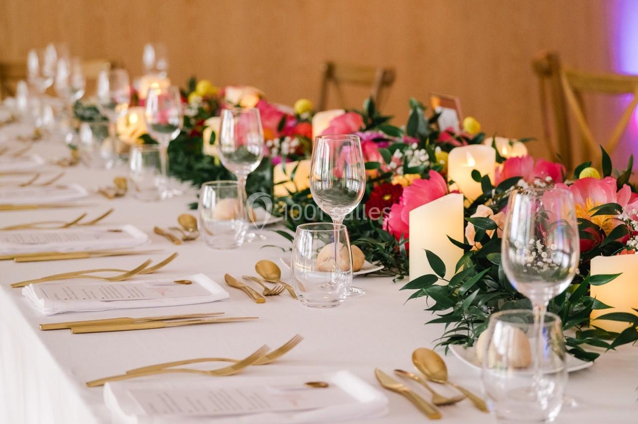 Table élégamment dressée avec nappes blanches, couverts dorés, verres, bougies et décorations florales colorées.
