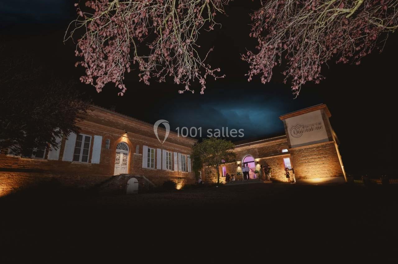 Bâtiment en briques éclairé de nuit, entouré d'arbres, avec des lumières mettant en valeur l'architecture.