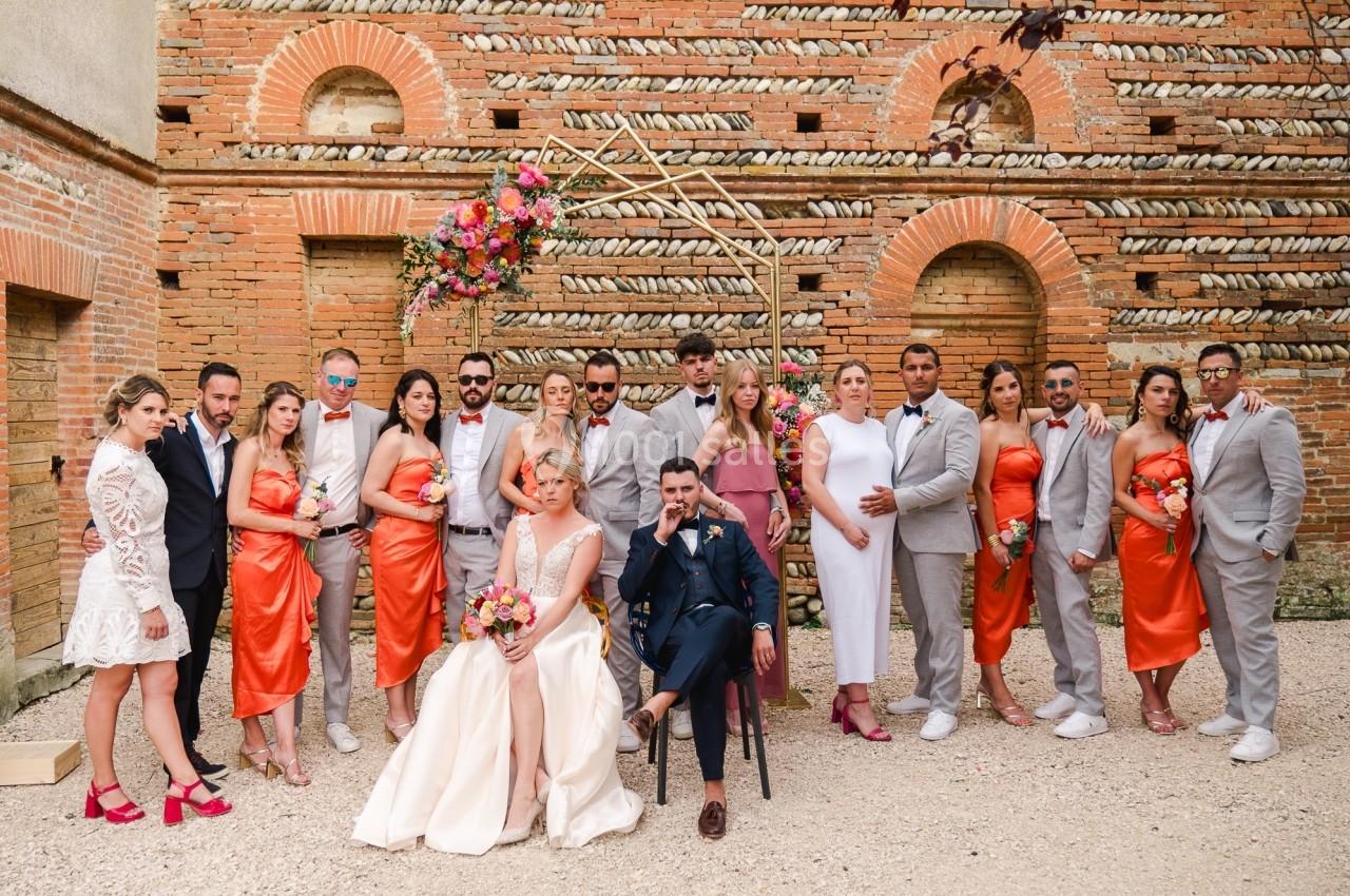 Un couple de mariés entouré de leurs invités en tenue élégante, posant devant un mur en briques décoré de fleurs.