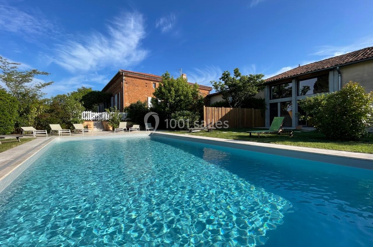 Piscine extérieure entourée de chaises longues, jardin arboré et maisons en arrière-plan sous un ciel bleu.