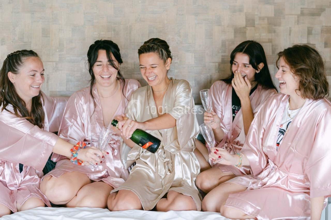 Cinq femmes en peignoirs satinés rient et trinquent avec une bouteille de champagne sur un lit.