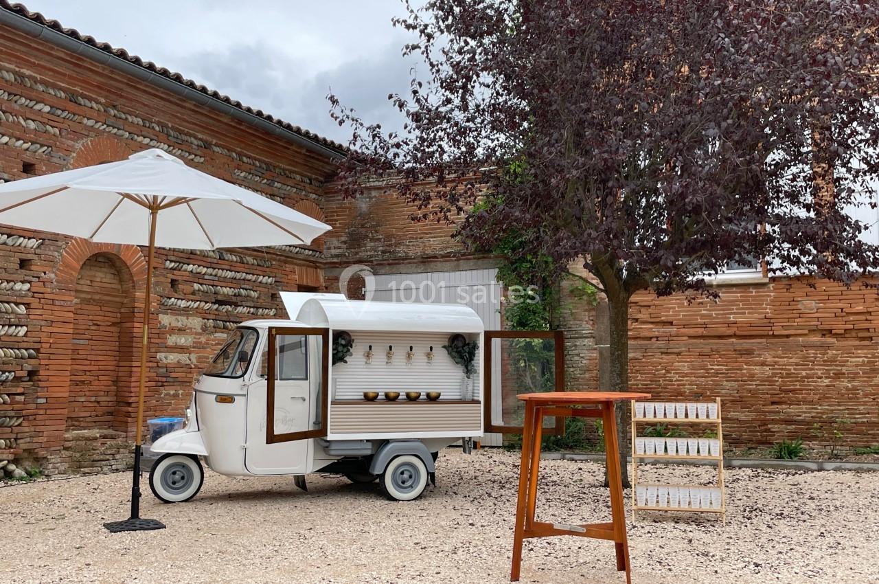 Triporteur blanc aménagé en bar mobile, stationné dans une cour pavée avec un parasol, un arbre et une table haute.