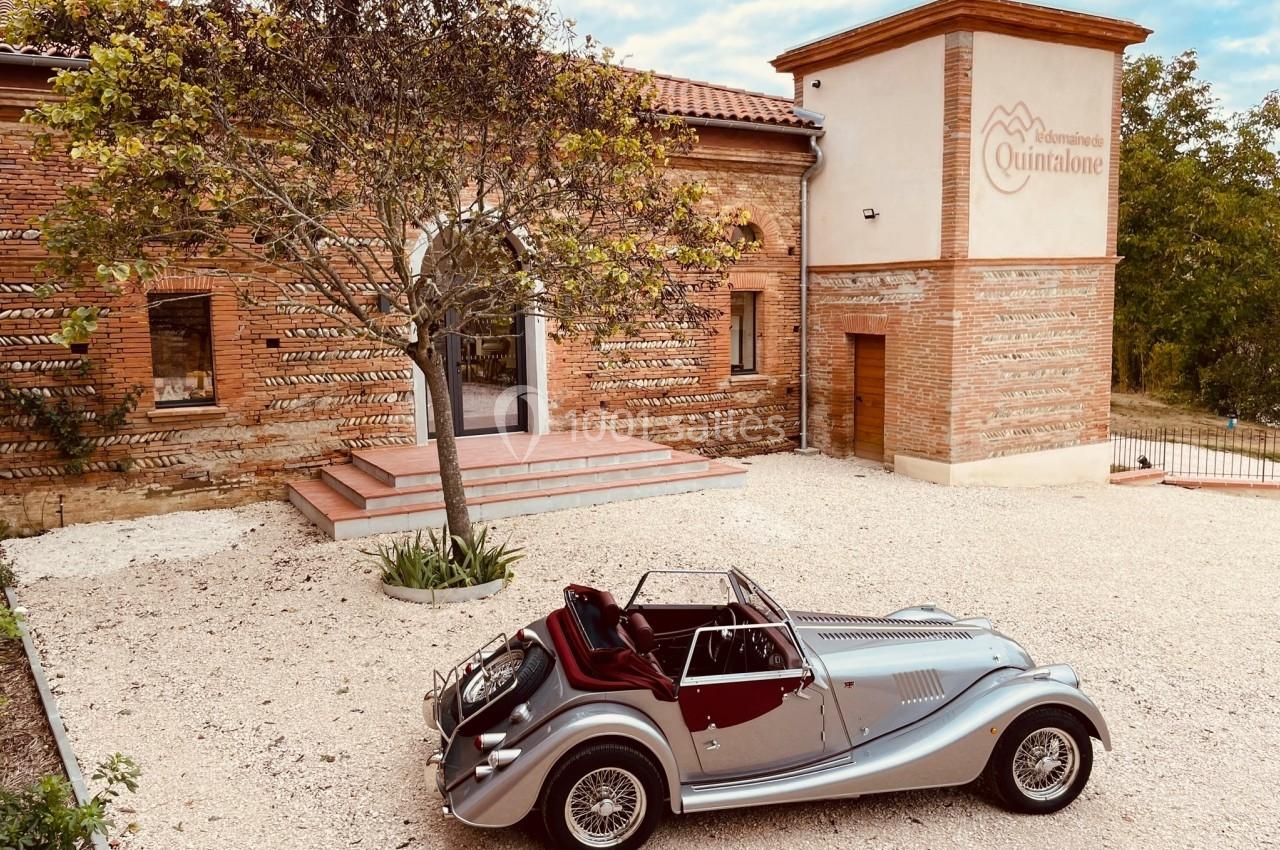Voiture ancienne argentée garée devant un bâtiment en briques avec un arbre au centre de la cour.