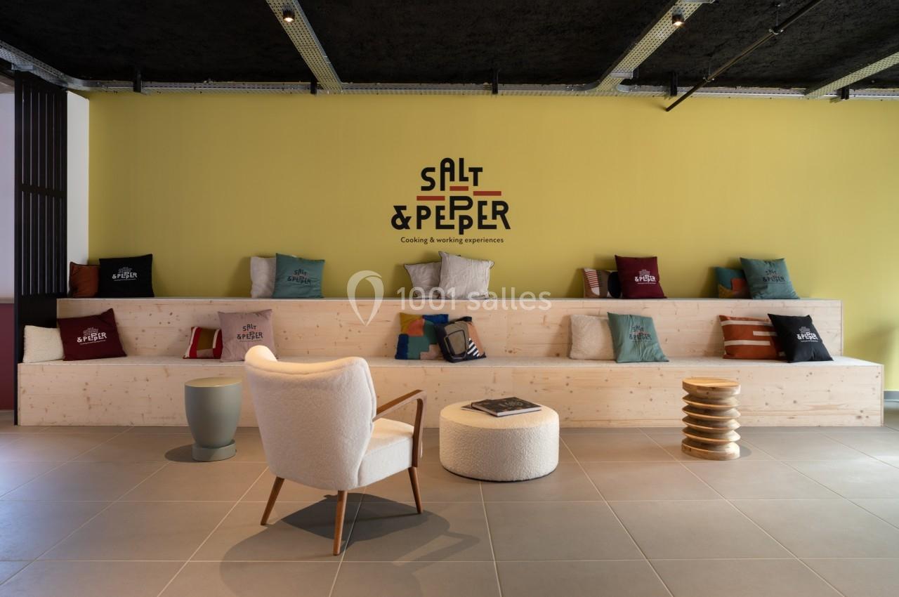 Espace lounge avec gradins en bois, coussins colorés, fauteuil et pouf, mur jaune avec logo ’Salt & Pepper’.