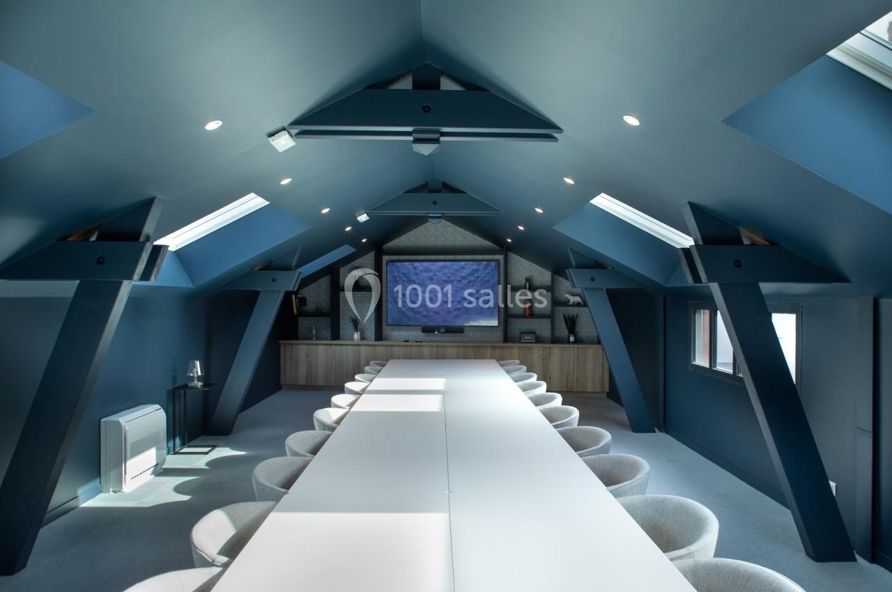 Salle de réunion moderne sous combles, avec une longue table blanche, chaises grises et écran mural au fond.