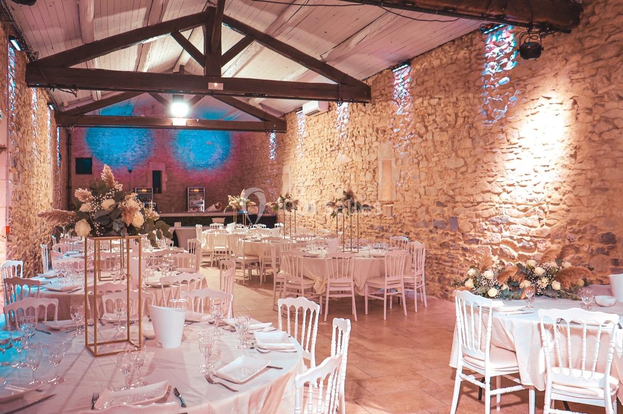 Salle de réception en pierre avec tables décorées de fleurs et chaises blanches, éclairée par une lumière tamisée.