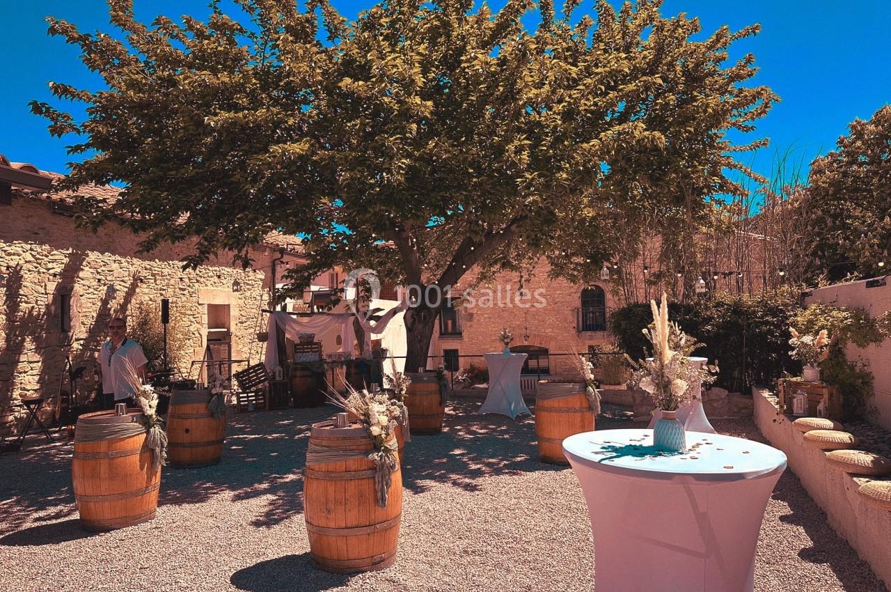 Cour extérieure ensoleillée avec des tonneaux en bois, un grand arbre central et des décorations pour un événement.