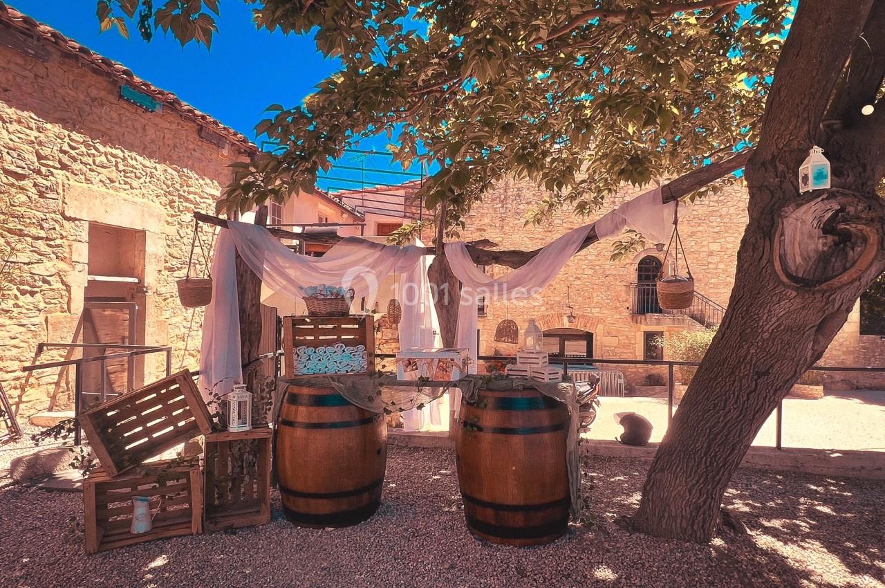 Décoration extérieure rustique avec tonneaux, caisses en bois et voilages sous un arbre dans une cour ensoleillée.