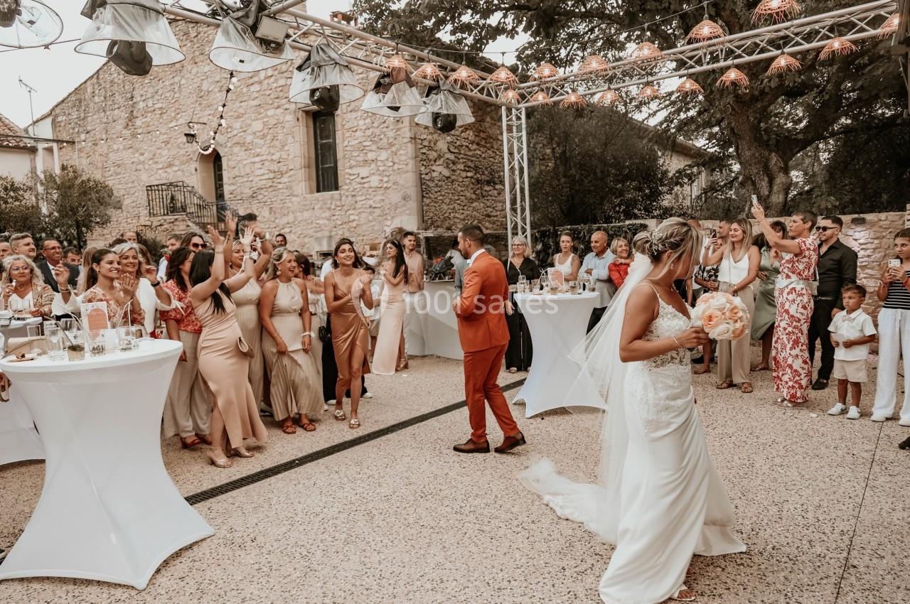 Un couple de mariés danse en extérieur devant des invités rassemblés autour de tables hautes.