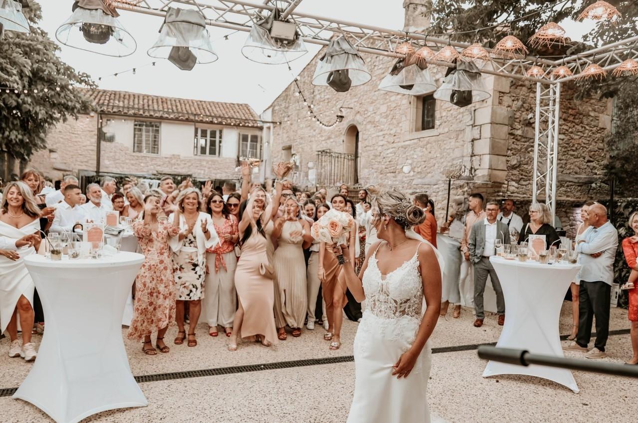 Une mariée lance son bouquet devant un groupe d'invités rassemblés dans une cour extérieure ensoleillée.