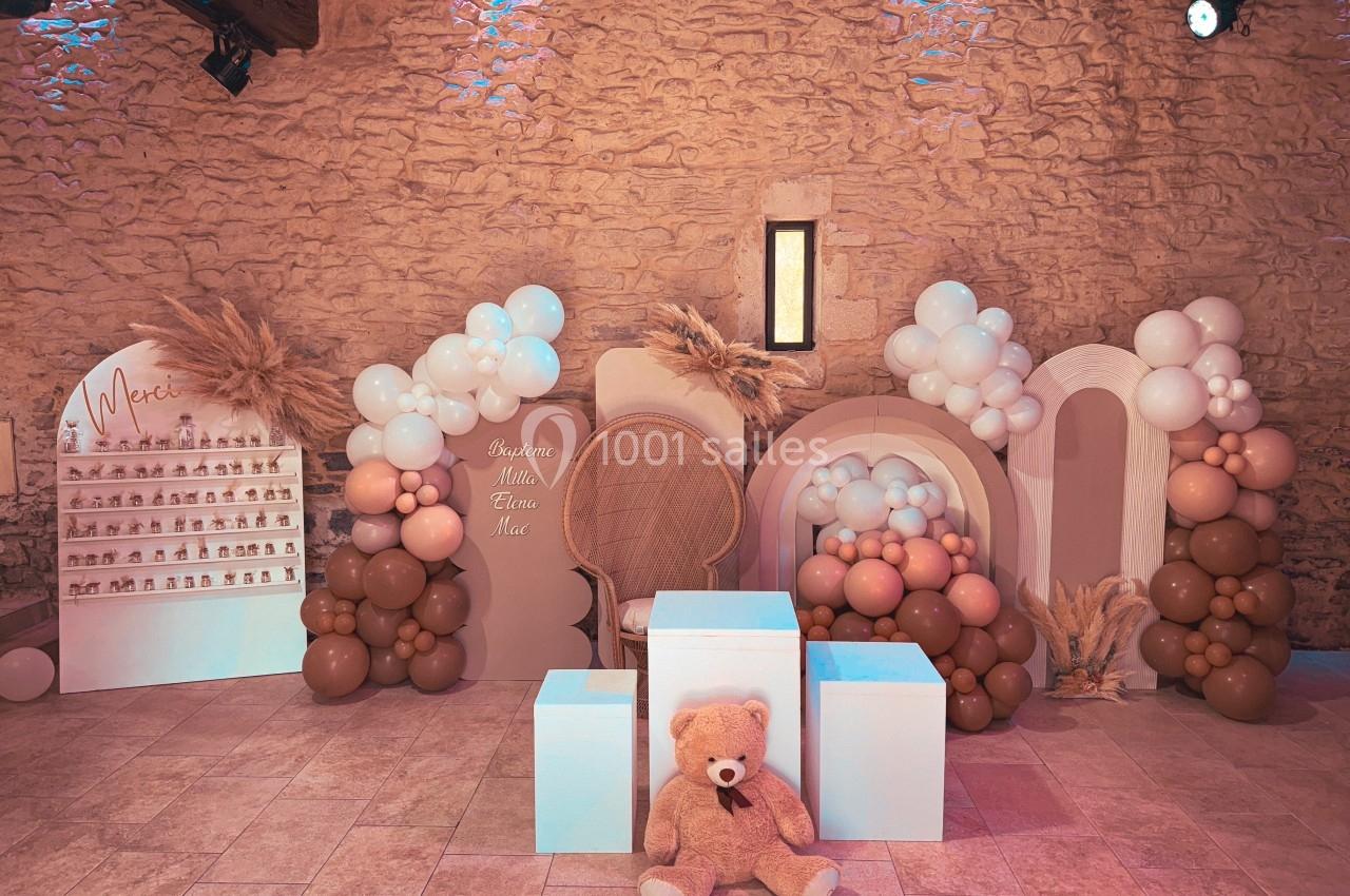 Décor de fête avec ballons beige et blanc, murs en pierre, présentoirs et un ours en peluche au centre.