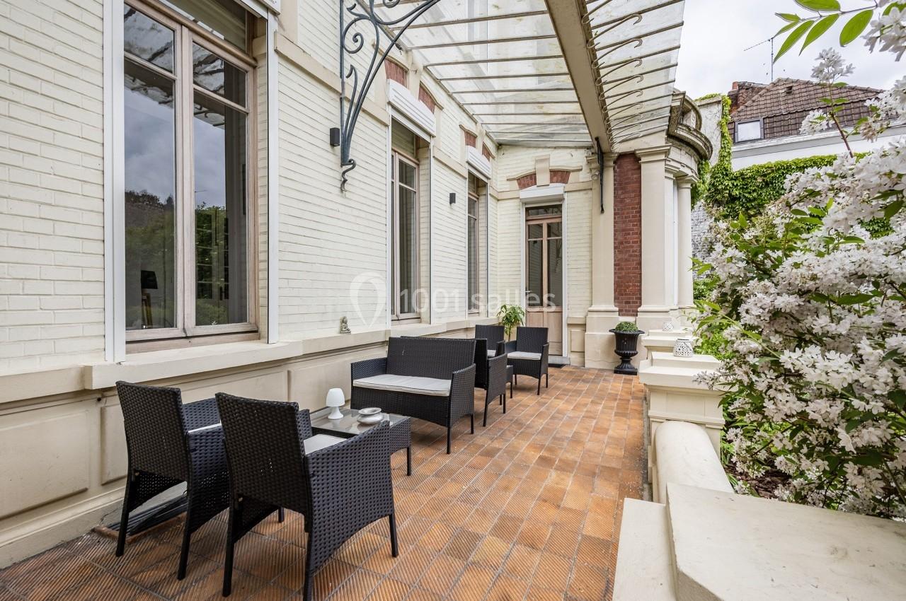 Terrasse en briques avec mobilier en rotin noir, adossée à une façade claire et entourée de végétation fleurie.