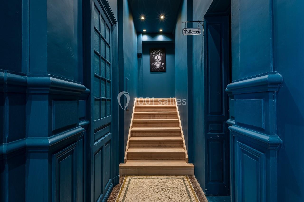 Escalier en bois éclairé menant à un étage, encadré de murs bleu foncé avec un tableau et une enseigne ’Toilettes’.