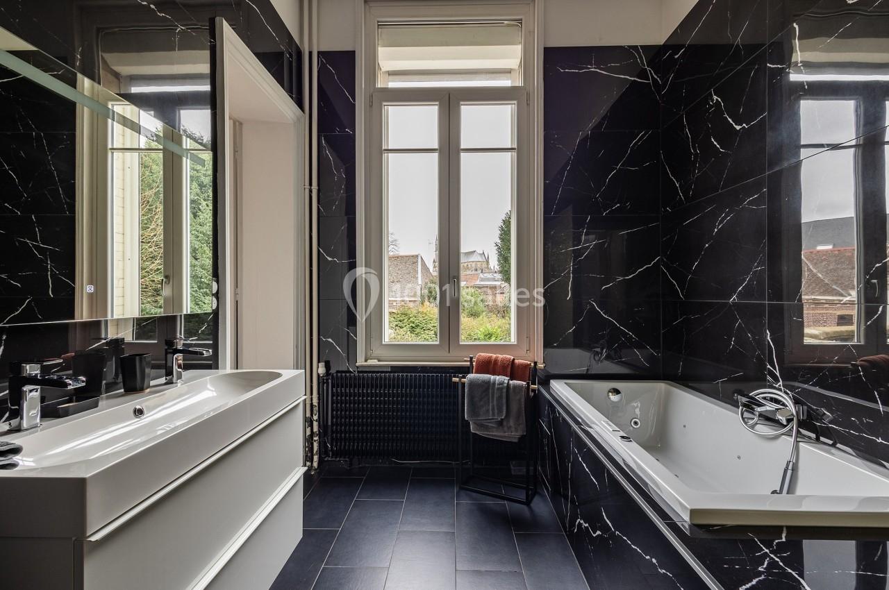 Salle de bain moderne avec murs noirs effet marbre, baignoire, lavabo blanc et grande fenêtre donnant sur un jardin.