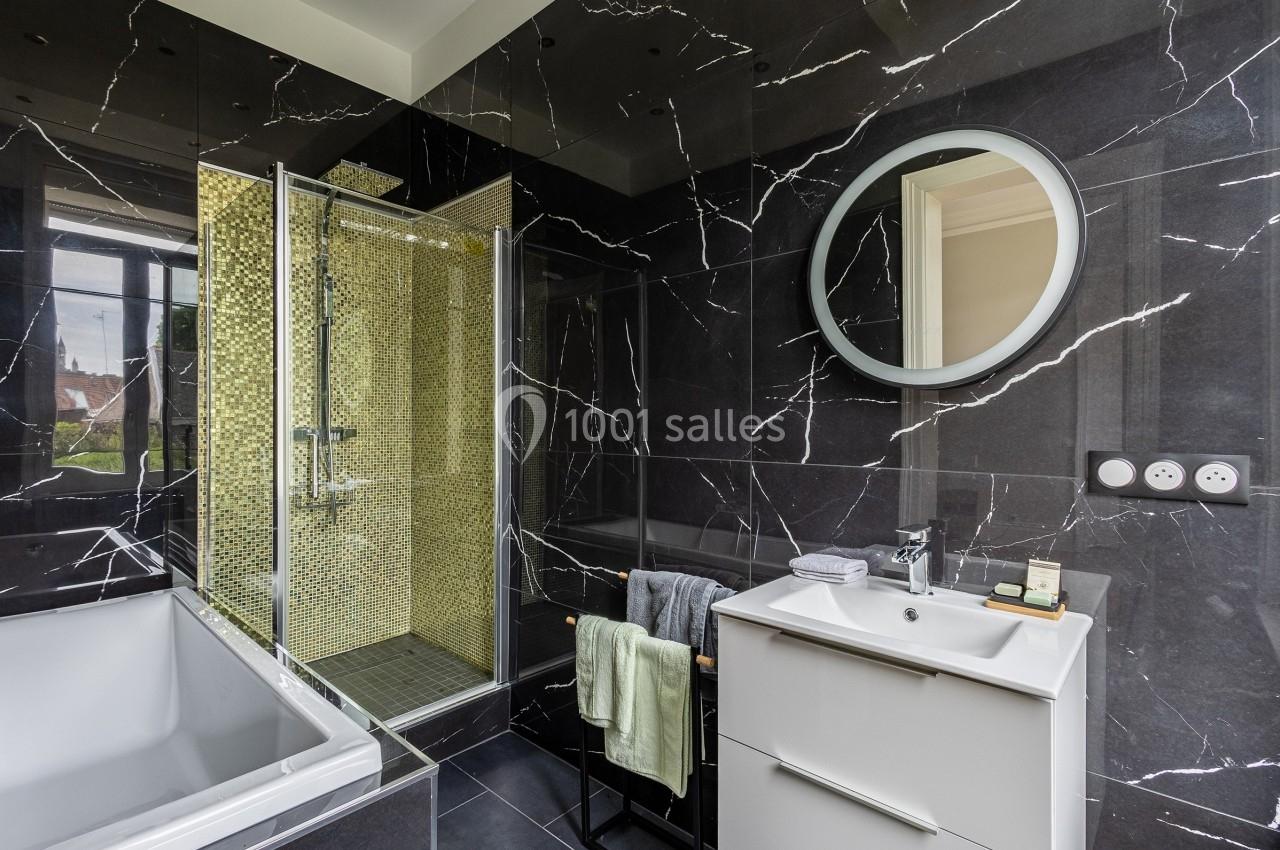Salle de bain moderne avec murs en marbre noir, douche à mosaïque dorée, baignoire blanche et meuble vasque blanc.