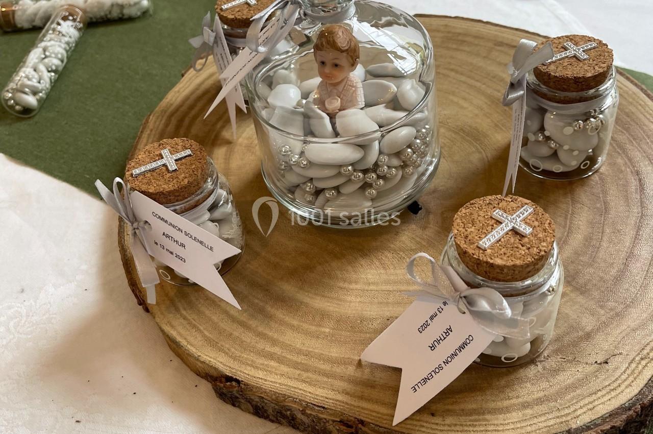 Petits pots de dragées et figurine sous cloche en décoration sur une rondelle de bois, disposés sur une table.