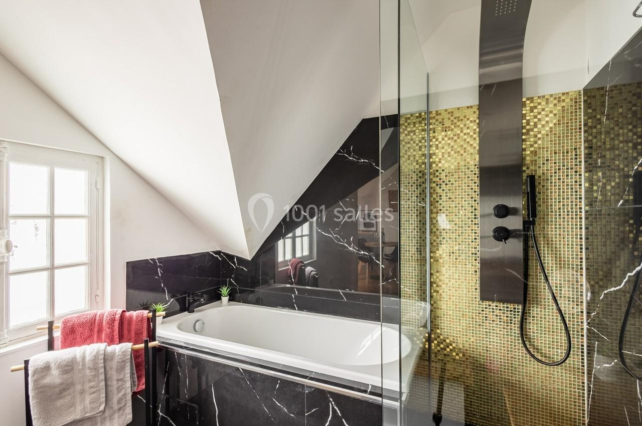 Salle de bain moderne avec baignoire encastrée, murs en marbre noir et douche à mosaïque dorée sous un toit mansardé.