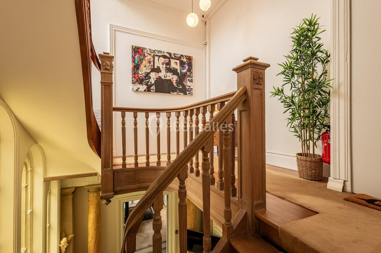 Escalier en bois avec rampe sculptée, plante en pot et tableau coloré accroché sur un mur blanc.
